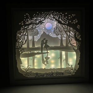 K&ouml;nnte beinhalten: Ein wei&szlig; gerahmtes, beleuchtetes Papier-Schnittkunstwerk, das ein Paar zeigt, das in einem Wald mit Mond und Sternen am Himmel steht. Der Text "June 2nd, 2018" befindet sich am unteren Rand des Bildes.