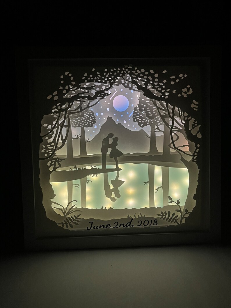 K&ouml;nnte beinhalten: Ein wei&szlig; gerahmtes, beleuchtetes Papier-Schnittkunstwerk, das ein Paar zeigt, das in einem Wald mit Mond und Sternen am Himmel steht. Der Text "June 2nd, 2018" befindet sich am unteren Rand des Bildes.