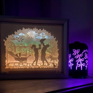 K&ouml;nnte beinhalten: Ein wei&szlig;er Rahmen mit einem Licht im Inneren, das eine Silhouette einer Familie in einem Park darstellt. Das Licht scheint durch ausgeschnittene Papierdesigns und erzeugt einen Schatteneffekt. Ein violettes Licht beleuchtet einen schwarzen Diffusor mit einem floralen Design im Hintergrund.
