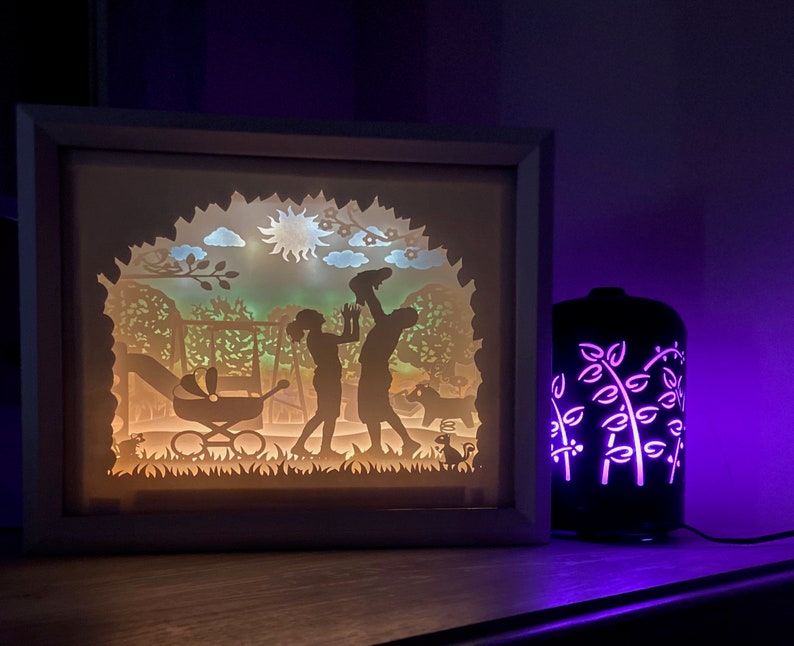 K&ouml;nnte beinhalten: Ein wei&szlig;er Rahmen mit einem Licht im Inneren, das eine Silhouette einer Familie in einem Park darstellt. Das Licht scheint durch ausgeschnittene Papierdesigns und erzeugt einen Schatteneffekt. Ein violettes Licht beleuchtet einen schwarzen Diffusor mit einem floralen Design im Hintergrund.