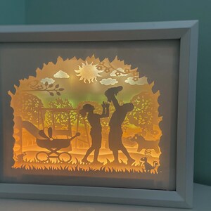 K&ouml;nnte beinhalten: Eine wei&szlig;e, gerahmte Papier-Ausschneide-Leuchtbox mit einer Silhouette einer Familie in einem Park. Die Familie besteht aus zwei Erwachsenen und einem Baby in einem Kinderwagen. Die Erwachsenen halten sich an den H&auml;nden und der Mann h&auml;lt das Baby in die Luft. Der Hintergrund ist eine Parkszene mit B&auml;umen, einer Schaukel und einer Sonne.