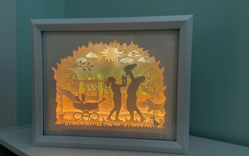 K&ouml;nnte beinhalten: Eine wei&szlig;e, gerahmte Papier-Ausschneide-Leuchtbox mit einer Silhouette einer Familie in einem Park. Die Familie besteht aus zwei Erwachsenen und einem Baby in einem Kinderwagen. Die Erwachsenen halten sich an den H&auml;nden und der Mann h&auml;lt das Baby in die Luft. Der Hintergrund ist eine Parkszene mit B&auml;umen, einer Schaukel und einer Sonne.