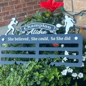 May include: A black metal medal display with the text "Champion Alisha She believed, She could, So She did". The display has six slots for medals and is decorated with two white silhouettes of tennis players.