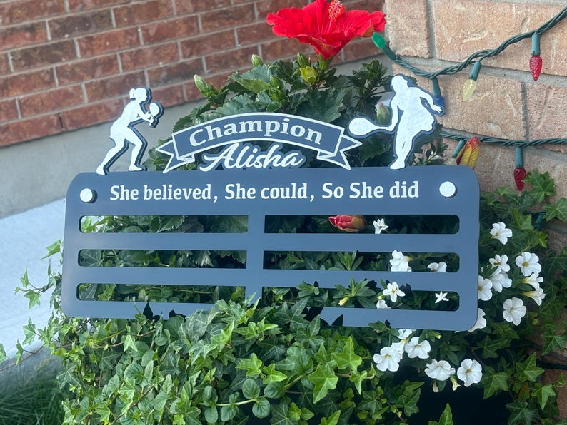 May include: A black metal medal display with the text "Champion Alisha She believed, She could, So She did". The display has six slots for medals and is decorated with two white silhouettes of tennis players.