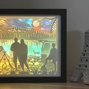 May include: A rectangular black-framed shadow box featuring a backlit layered paper cutout scene of a camping site with two people standing, a tent, a campfire, a vehicle, a deer, and a forest under a moonlit sky, next to a white Eiffel Tower figurine.