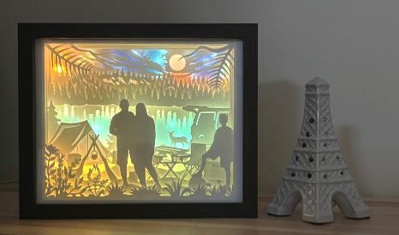 May include: A rectangular black-framed shadow box featuring a backlit layered paper cutout scene of a camping site with two people standing, a tent, a campfire, a vehicle, a deer, and a forest under a moonlit sky, next to a white Eiffel Tower figurine.