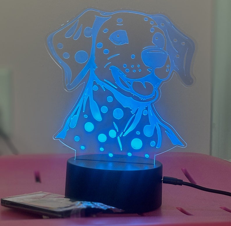 May include: A blue night light shaped like a Dalmatian dog with black spots. The light is on and illuminating the dog's face.