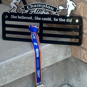 May include: A black metal medal display with the words "Champion Alisha" and "She believed, She could, So She did". A blue and orange ribbon with a silver medal is hanging from the display.