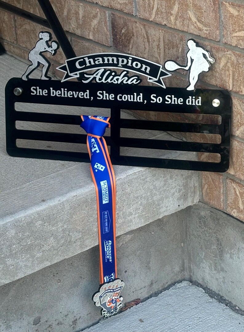 May include: A black metal medal display with the words "Champion Alisha" and "She believed, She could, So She did". A blue and orange ribbon with a silver medal is hanging from the display.