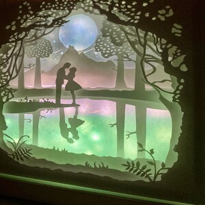 K&ouml;nnte beinhalten: Ein Papier-Schnittbild, das ein Paar zeigt, das an einem See in einem Wald steht. Die Szene wird von hinten beleuchtet, wodurch ein leuchtender Effekt entsteht. Das Paar ist vor einem Vollmond und einem Berg in der Ferne silhouettiert.
