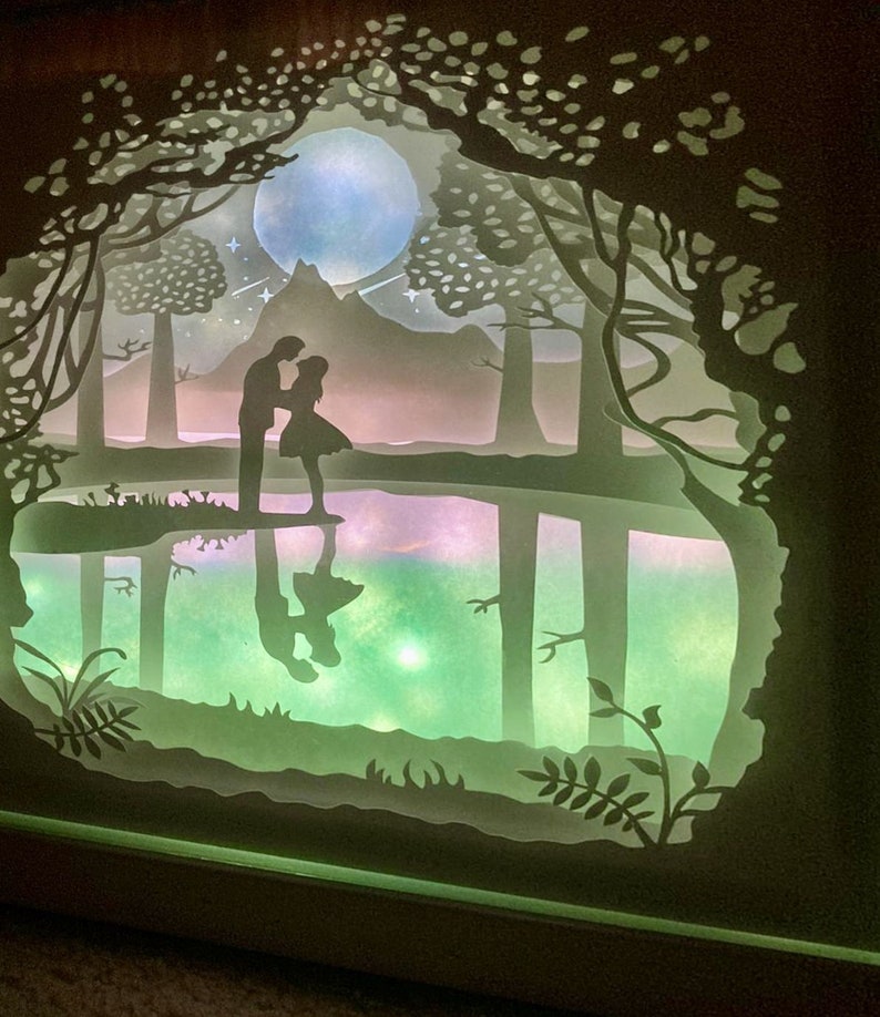 K&ouml;nnte beinhalten: Ein Papier-Schnittbild, das ein Paar zeigt, das an einem See in einem Wald steht. Die Szene wird von hinten beleuchtet, wodurch ein leuchtender Effekt entsteht. Das Paar ist vor einem Vollmond und einem Berg in der Ferne silhouettiert.