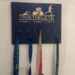 K&ouml;nnte beinhalten: Eine blaue Holzplakette mit den Worten "TRIATHELETE" und "★ SWIM ★ BIKE ★ RUN ★" in Wei&szlig;. An der Plakette befinden sich acht Haken, an denen drei Medaillen h&auml;ngen. Die Medaillen sind blau, rot und gelb.