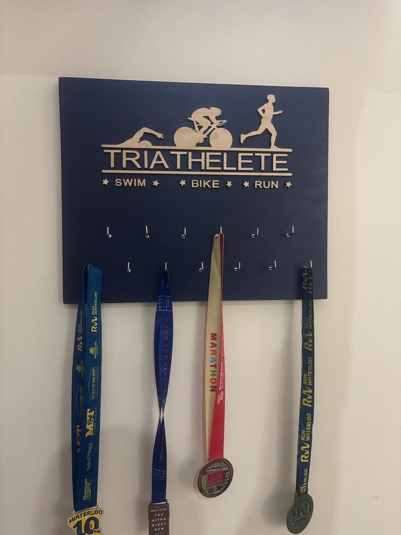 K&ouml;nnte beinhalten: Eine blaue Holzplakette mit den Worten "TRIATHELETE" und "★ SWIM ★ BIKE ★ RUN ★" in Wei&szlig;. An der Plakette befinden sich acht Haken, an denen drei Medaillen h&auml;ngen. Die Medaillen sind blau, rot und gelb.