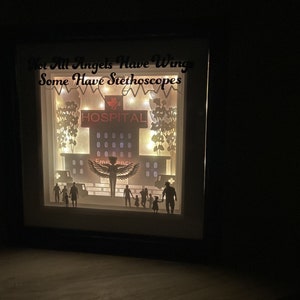 May include: A black framed light-up box with a white background. The box features a paper cut-out design of a hospital building with the word "HOSPITAL" above the entrance. The text "Not All Angels Have Wings Some Have Stethoscopes" is written above the building. The box is lit up with white lights.