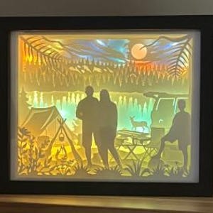 May include: A rectangular shadow box with a black frame featuring a multi-layered paper cut-out scene of a campsite at sunset. The backlit display shows silhouettes of three people, a tent, a vehicle, a deer, and trees against a glowing sky.
