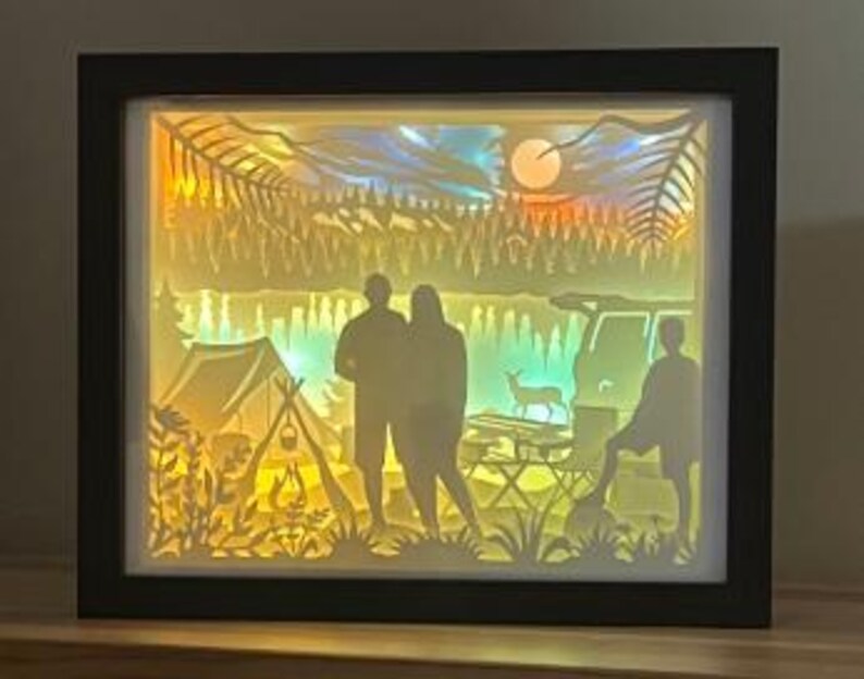 May include: A rectangular shadow box with a black frame featuring a multi-layered paper cut-out scene of a campsite at sunset. The backlit display shows silhouettes of three people, a tent, a vehicle, a deer, and trees against a glowing sky.