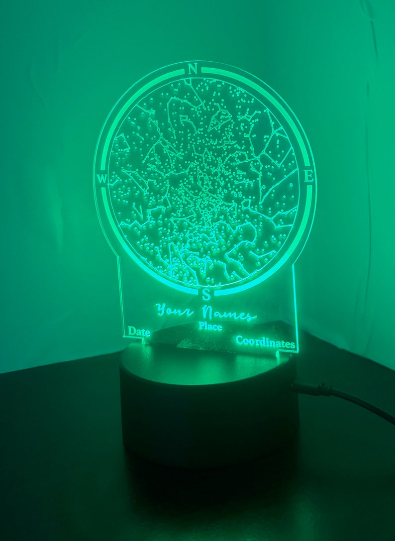 Op de afbeelding: Een groen nachtlampje met een cirkelvormig ontwerp dat een sterrenkaart voorstelt. Het lampje heeft een zwarte voet en bevat de tekst "Your Names", "Date", "Place" en "Coordinates".