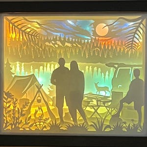 May include: A rectangular shadow box with a dark frame containing a multi-layered paper cutout scene of a camping trip. The backlit display features silhouettes of two adults and a child, a tent, a campfire, a vehicle, a deer, and a forest.