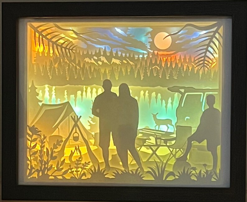 May include: A rectangular shadow box with a dark frame containing a multi-layered paper cutout scene of a camping trip. The backlit display features silhouettes of two adults and a child, a tent, a campfire, a vehicle, a deer, and a forest.