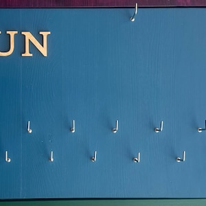 May include: A blue wooden board with the word "RUN" in gold letters. The board has 12 hooks for hanging medals or other items.