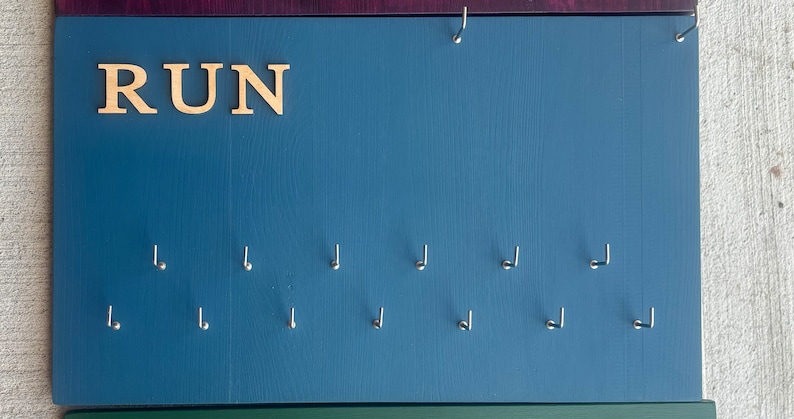 May include: A blue wooden board with the word "RUN" in gold letters. The board has 12 hooks for hanging medals or other items.