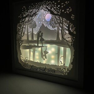 K&ouml;nnte beinhalten: Ein wei&szlig;es Papierkunstwerk mit einem Paar, das in einer Waldszene silhouettiert ist. Die Szene enth&auml;lt einen Mond, Sterne und einen Berg. Das Datum "June 2nd, 2018" ist unten geschrieben.