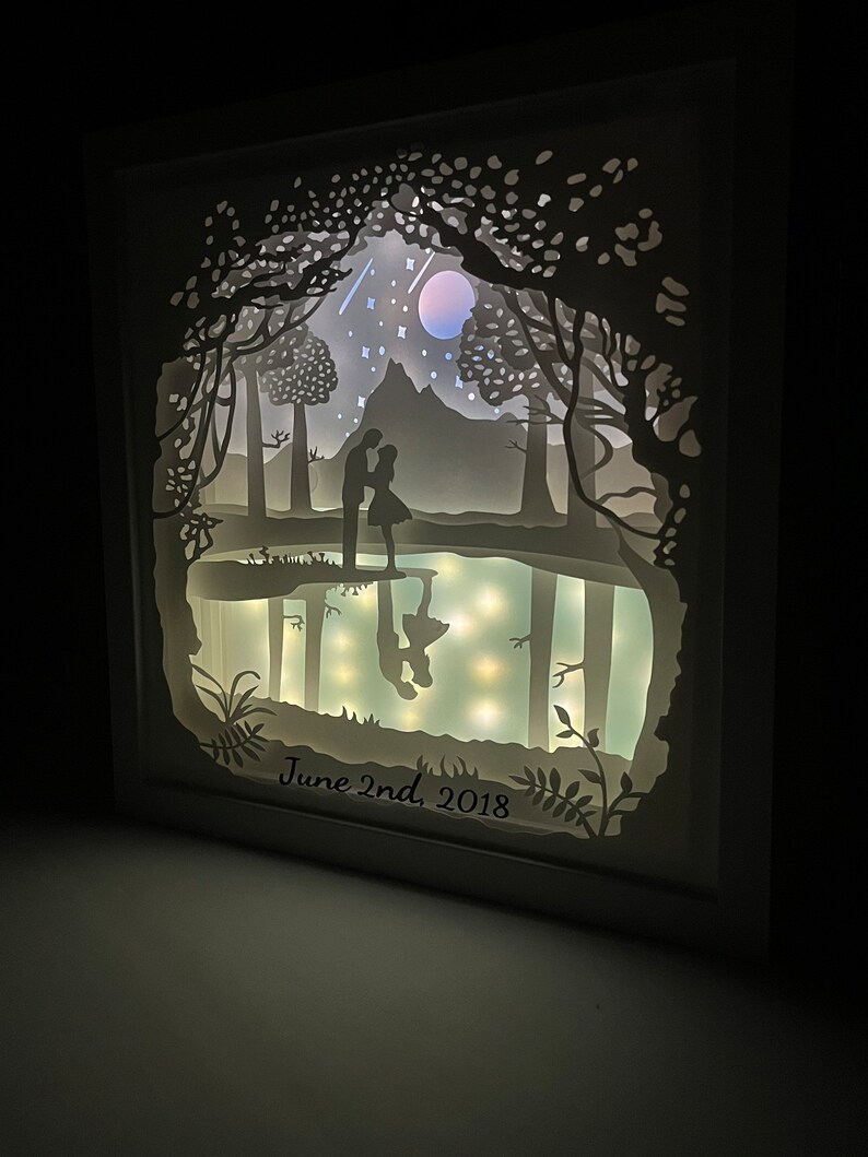 K&ouml;nnte beinhalten: Ein wei&szlig;es Papierkunstwerk mit einem Paar, das in einer Waldszene silhouettiert ist. Die Szene enth&auml;lt einen Mond, Sterne und einen Berg. Das Datum "June 2nd, 2018" ist unten geschrieben.