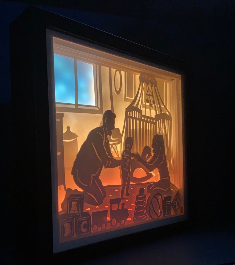 Op de afbeelding: Een schaduwdoos van papier met een silhouet van een gezin dat met speelgoed speelt. Het gezin bestaat uit een vader, een moeder en een kind. Het speelgoed omvat een trein, blokken en een bal. De schaduwdoos wordt van achteren verlicht, waardoor een warme gloed ontstaat.