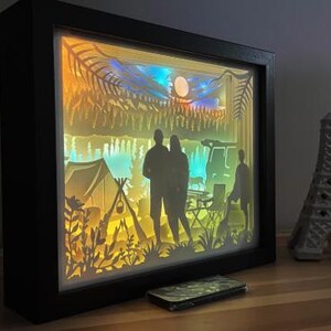May include: A rectangular black shadow box frame containing a multi-layered paper cutout scene of a camping trip. The backlit display features silhouettes of two people, a tent, a campfire, trees, and a vehicle against a gradient of yellow and blue.