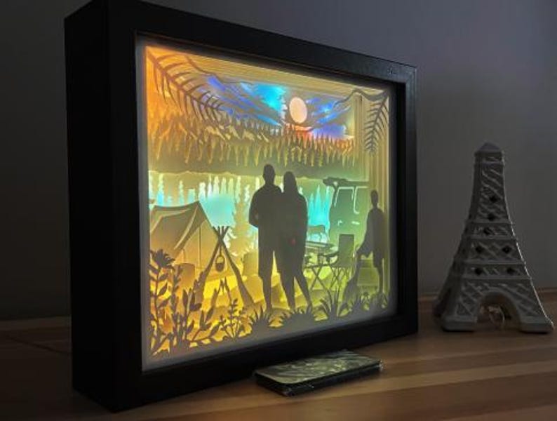 May include: A rectangular black shadow box frame containing a multi-layered paper cutout scene of a camping trip. The backlit display features silhouettes of two people, a tent, a campfire, trees, and a vehicle against a gradient of yellow and blue.
