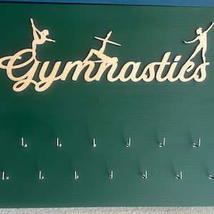 Puede incluir: Un tablero de madera verde con la palabra "Gimnasia" recortada en madera y dos siluetas de gimnastas. El tablero tiene 10 ganchos plateados para colgar medallas o cintas.