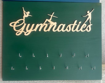 May include: A green wooden board with the word "Gymnastics" cut out in wood and two gymnast silhouettes. The board has 10 silver hooks for hanging medals or ribbons.