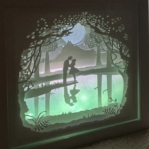 K&ouml;nnte beinhalten: Ein wei&szlig;es Papier-Schnittbild in einem Rahmen mit einem hellgr&uuml;nen Leuchten. Die Kunst zeigt ein Paar, das in einem Wald mit einem Vollmond und einem Berg im Hintergrund steht.
