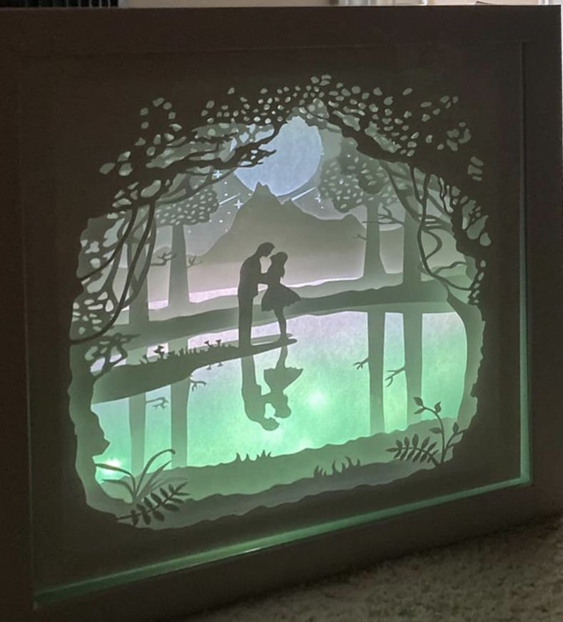 K&ouml;nnte beinhalten: Ein wei&szlig;es Papier-Schnittbild in einem Rahmen mit einem hellgr&uuml;nen Leuchten. Die Kunst zeigt ein Paar, das in einem Wald mit einem Vollmond und einem Berg im Hintergrund steht.
