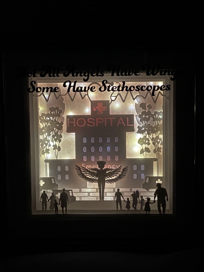 May include: A papercraft shadow box with a white background and a silhouette of an angel with wings, a hospital building, and people walking. The text "Not all angels have wings, some have stethoscopes" is at the top of the box.