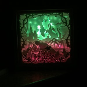 May include: A paper cut-out light-up scene with a green and pink glow. The scene features a castle, a dragon, a fairy, and other characters. The scene is framed in a white frame.