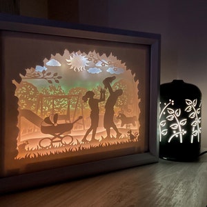 K&ouml;nnte beinhalten: Ein wei&szlig;er Rahmen mit einem Licht im Inneren, der eine Familie aus drei Silhouetten in einer Parklandschaft zeigt. Die Szene beinhaltet eine Schaukel, einen Kinderwagen und eine Sonne. Der Rahmen steht auf einer Holzoberfl&auml;che neben einer schwarzen Lampe mit einem wei&szlig;en Blumendesign.