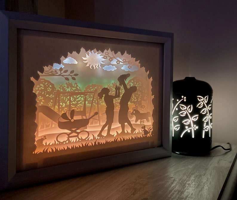 K&ouml;nnte beinhalten: Ein wei&szlig;er Rahmen mit einem Licht im Inneren, der eine Familie aus drei Silhouetten in einer Parklandschaft zeigt. Die Szene beinhaltet eine Schaukel, einen Kinderwagen und eine Sonne. Der Rahmen steht auf einer Holzoberfl&auml;che neben einer schwarzen Lampe mit einem wei&szlig;en Blumendesign.