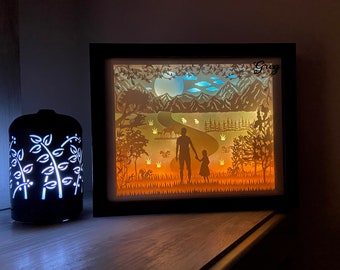 Puede incluir: Una pieza de arte de papel iluminada con marco negro con un diseño recortado de un padre y una hija caminando por una escena de bosque. La escena incluye un río, árboles, montañas y una luna llena. El nombre "Greg" está escrito en la esquina superior derecha del marco.