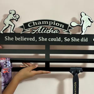 Puede incluir: Expositor de medallas negro con letras plateadas que dicen "Champion Alisha She believed, She could, So She did". El expositor tiene dos siluetas plateadas de jugadores de tenis con raquetas. Dos medallas cuelgan del expositor.