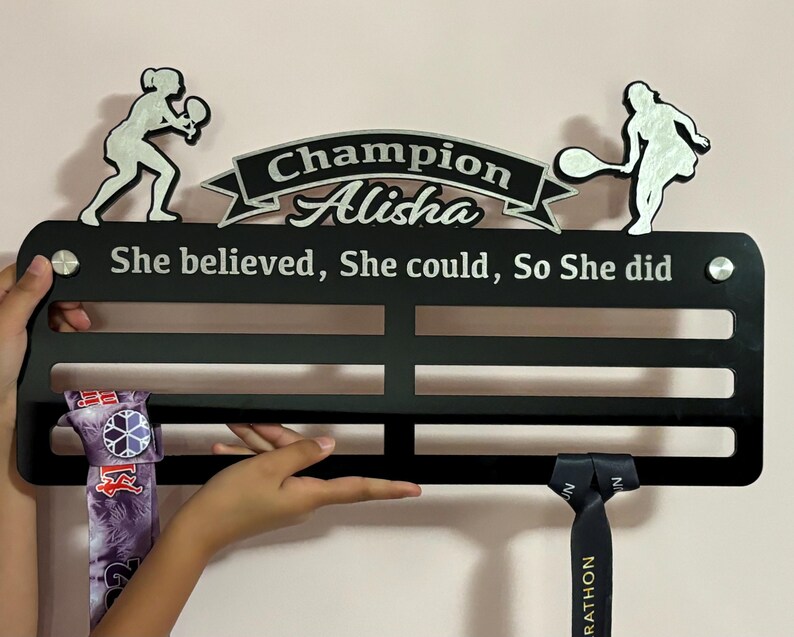 Puede incluir: Expositor de medallas negro con letras plateadas que dicen "Champion Alisha She believed, She could, So She did". El expositor tiene dos siluetas plateadas de jugadores de tenis con raquetas. Dos medallas cuelgan del expositor.
