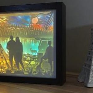 May include: A rectangular black-framed shadow box light featuring a multi-layered paper cutout scene of a camping site with a tent, a vehicle, a deer, and silhouettes of three people, illuminated by a warm, colourful gradient light.