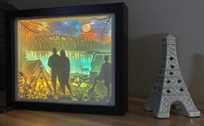May include: A rectangular black-framed shadow box light featuring a multi-layered paper cutout scene of a camping site with a tent, a vehicle, a deer, and silhouettes of three people, illuminated by a warm, colourful gradient light.