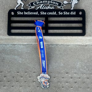 May include: Black metal medal display rack with the text "Champion Alisha She believed, She could, So She did". A blue and orange ribbon with a medal is hanging from the rack. The medal features a cartoon fox.