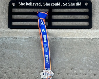 Puede incluir: Expositor de medallas de metal negro con el texto "Champion Alisha She believed, She could, So She did". Una cinta azul y naranja con una medalla cuelga del expositor. La medalla presenta un zorro de dibujos animados.