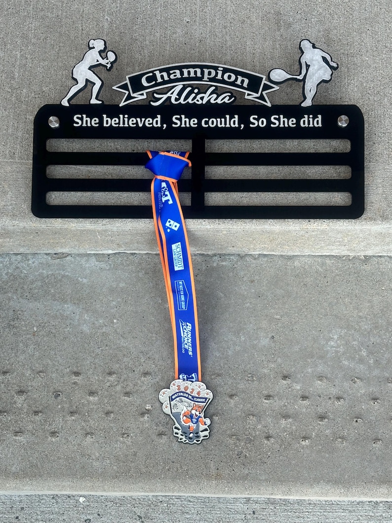 May include: Black metal medal display rack with the text "Champion Alisha She believed, She could, So She did". A blue and orange ribbon with a medal is hanging from the rack. The medal features a cartoon fox.