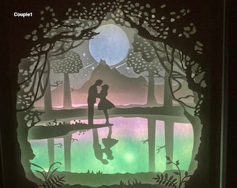 Könnte beinhalten: Ein weißes Papier-Schnittbild, das ein Paar zeigt, das in einem Wald mit Vollmond und Sternen am Himmel steht. Das Paar spiegelt sich in einem See im Vordergrund.
