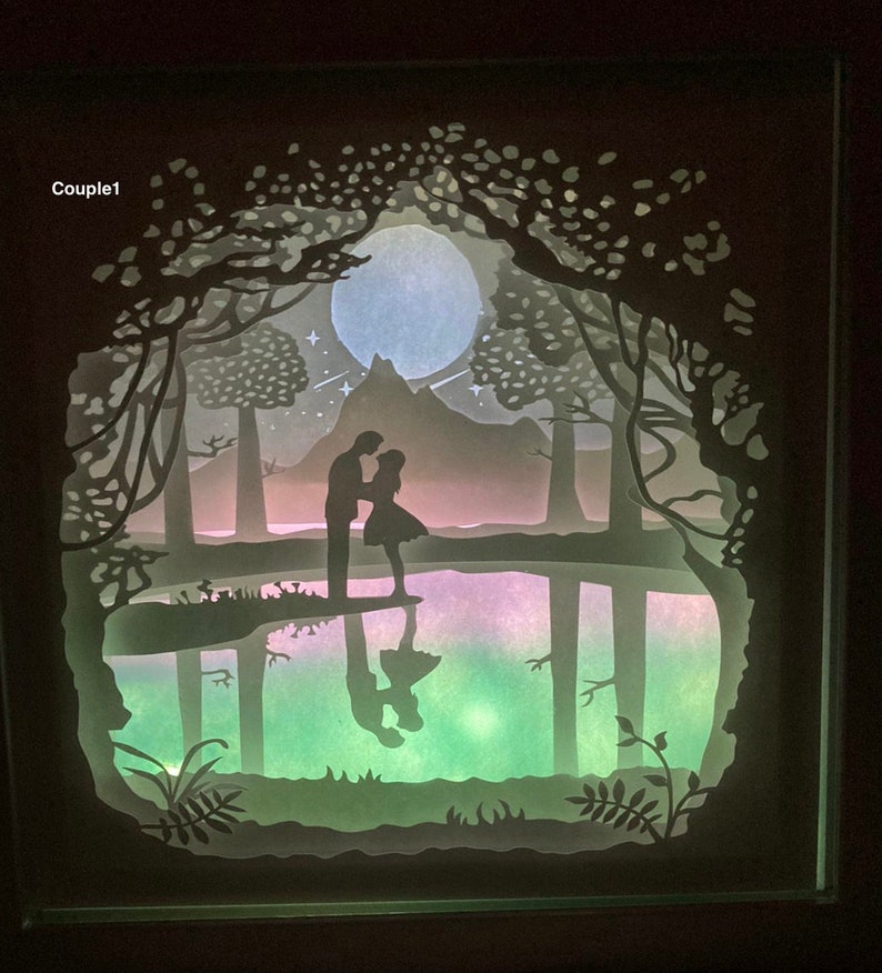 K&ouml;nnte beinhalten: Ein wei&szlig;es Papier-Schnittbild, das ein Paar zeigt, das in einem Wald mit Vollmond und Sternen am Himmel steht. Das Paar spiegelt sich in einem See im Vordergrund.