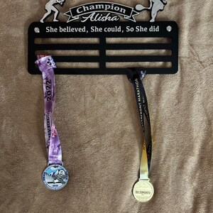 Puede incluir: Un soporte para medallas de metal negro con dos medallas colgando de &eacute;l. El soporte tiene el texto "Champion Alisha" y "She believed, She could, So She did". Las medallas son doradas y moradas con el texto "2022" y "2023" en ellas.