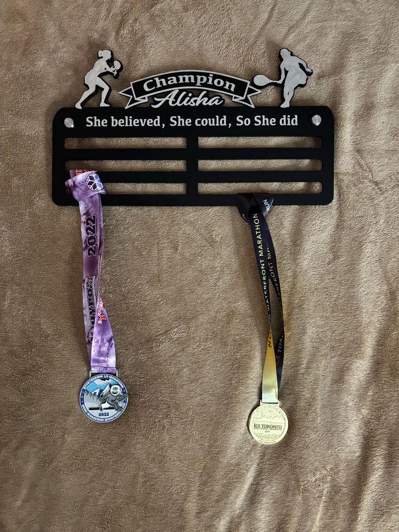Puede incluir: Un soporte para medallas de metal negro con dos medallas colgando de &eacute;l. El soporte tiene el texto "Champion Alisha" y "She believed, She could, So She did". Las medallas son doradas y moradas con el texto "2022" y "2023" en ellas.